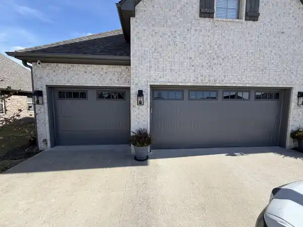 Grey raised-panel single and double garage doors with window tops on a pale tumbled-brick house