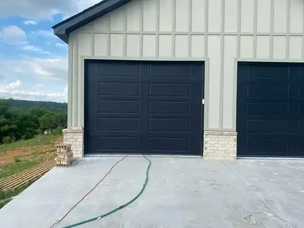 Two black raised-panel garage doors on a sage board-and-batten house with a hillside behind