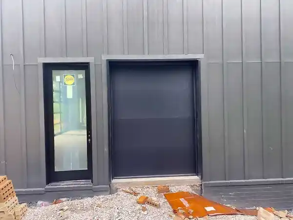 Flush black garage door beside a glass entry door in grey metal siding