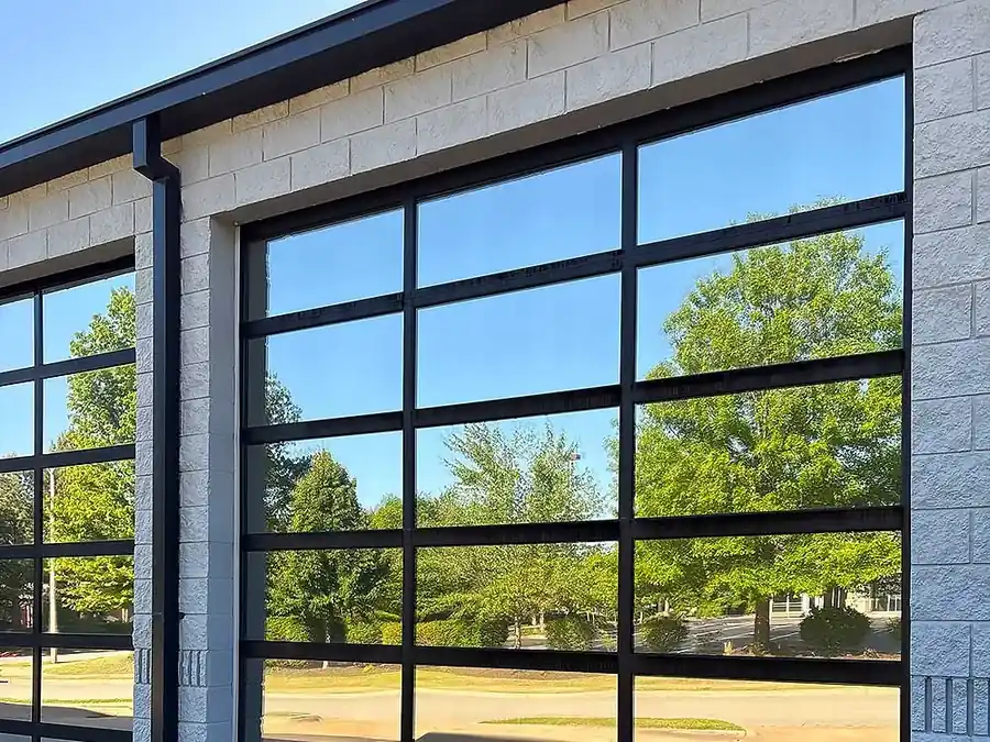 A black-framed full-view glass garage door installed by Elite, trees reflected in the panels