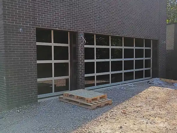 Black full-view garage doors set in dark brick with pallets on gravel in front